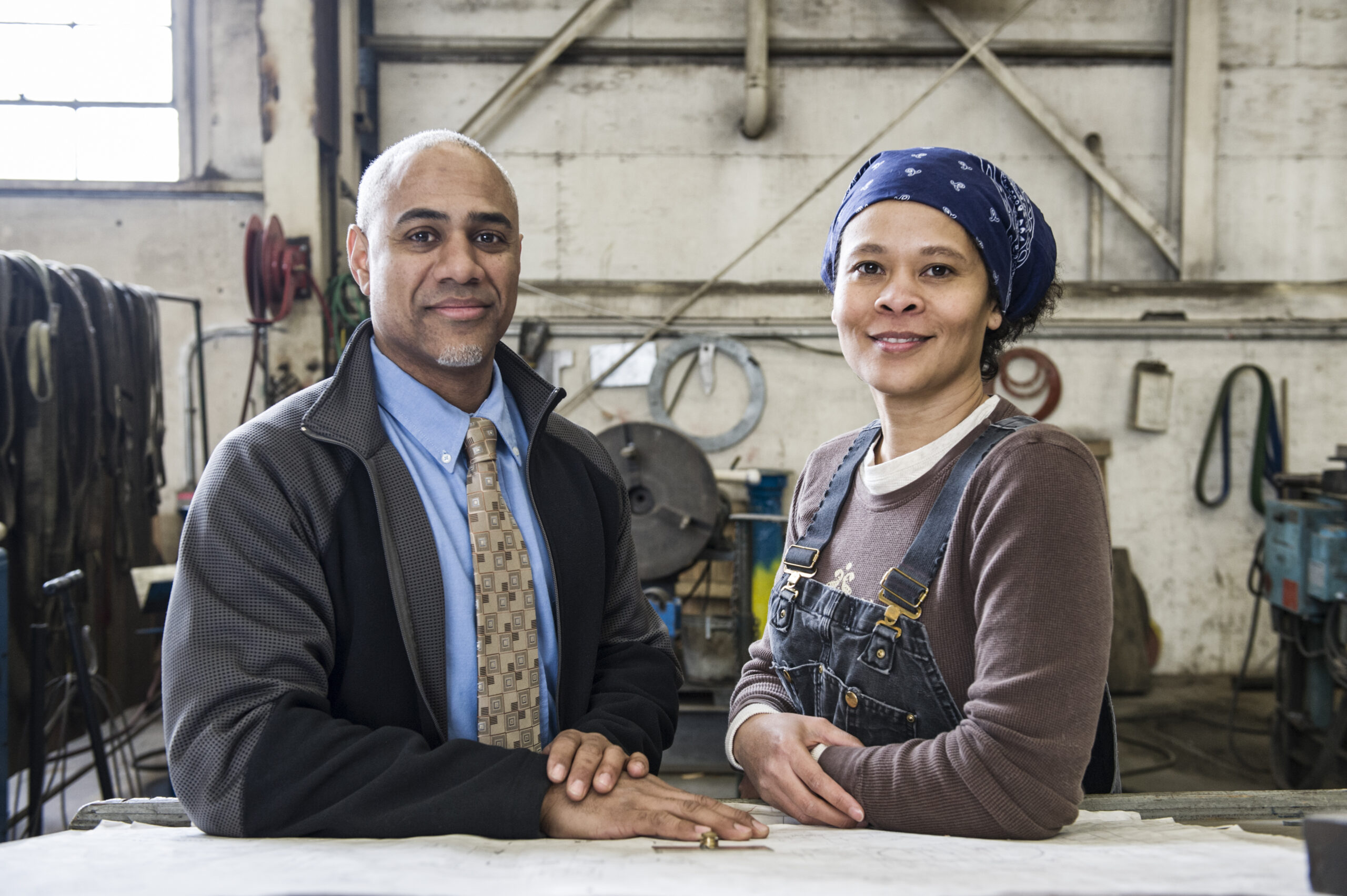 Black man owner of a sheet metal factoy and a black woman factory worker going over plans at a work station on the floor of the factory.